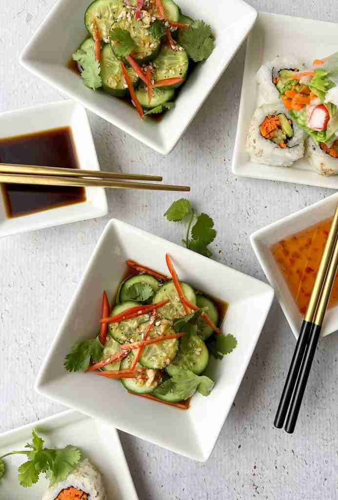 Asian cucumber salad in white bowls with plates of sushi and dipping sauces